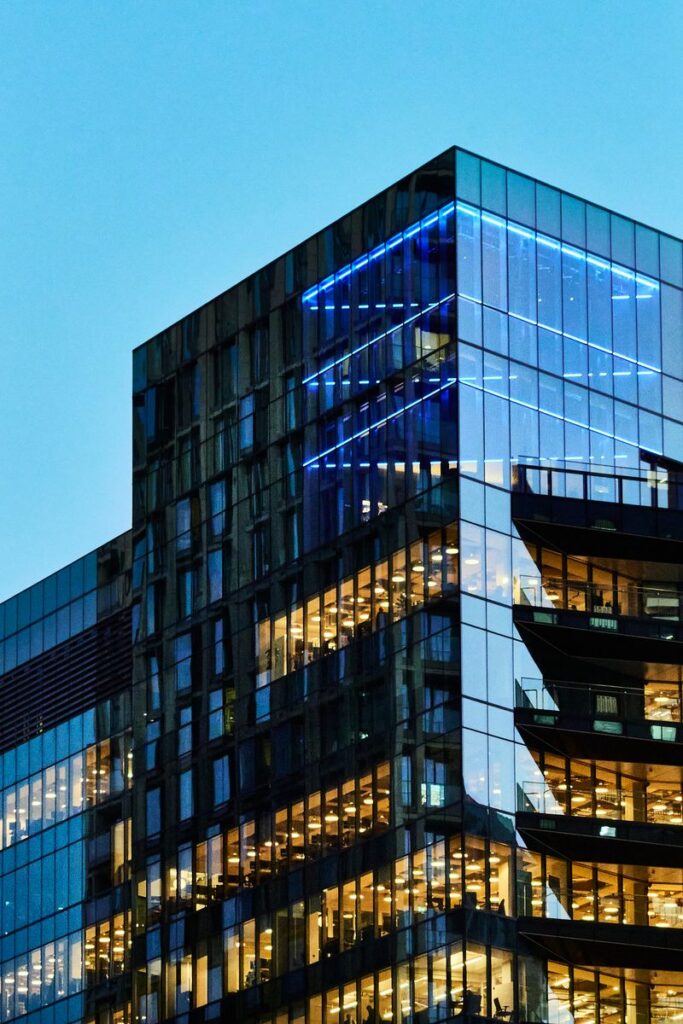 A sleek glass office building with illuminated interior visible against a twilight sky.